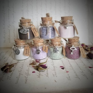 May include: Seven small glass jars with cork lids, each filled with a different colored sand and a small charm hanging from the lid. The jars are arranged in a pyramid shape on a book with text. The charms include an eye, a feather, a heart, a spiral, a four-leaf clover, a key, and a crystal.