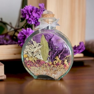 May include: A clear, round glass bottle filled with dried herbs and flowers, including purple petals, sage, and a bay leaf. The bottle has a cork stopper with a clear crystal on top and is tied with a white ribbon. The background includes purple flowers and a wooden box.