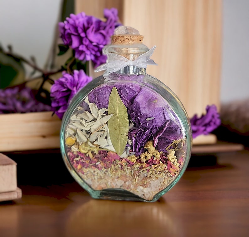 May include: A clear, round glass bottle filled with dried herbs and flowers, including purple petals, sage, and a bay leaf. The bottle has a cork stopper with a clear crystal on top and is tied with a white ribbon. The background includes purple flowers and a wooden box.