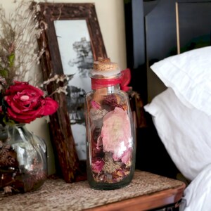 May include: A clear glass bottle filled with dried flowers and leaves, with a red ribbon tied around the neck. The bottle is sitting on a wooden nightstand next to a bed.