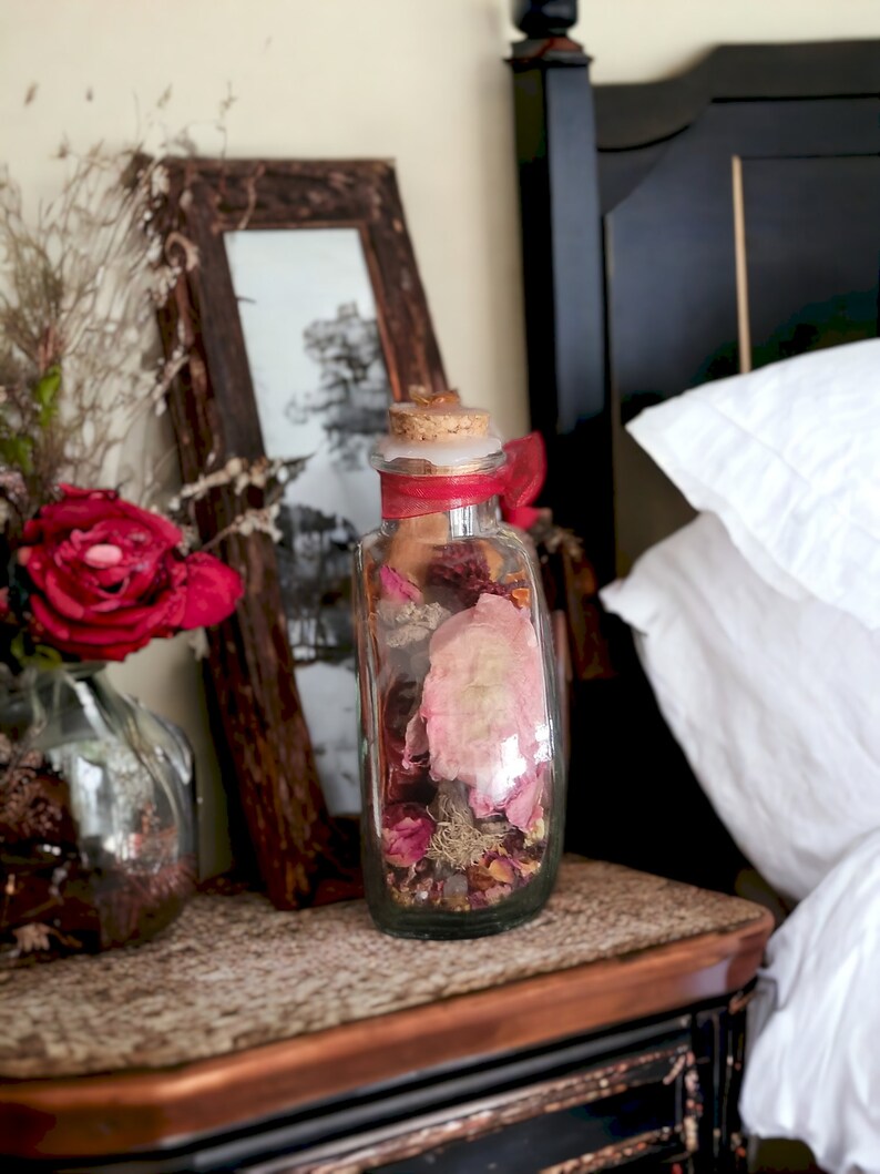 May include: A clear glass bottle filled with dried flowers and leaves, with a red ribbon tied around the neck. The bottle is sitting on a wooden nightstand next to a bed.