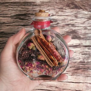May include: A clear glass jar with a cork stopper and a red ribbon tied around the neck. The jar is filled with dried herbs, spices, and rose petals. The jar is sitting on a wooden surface.