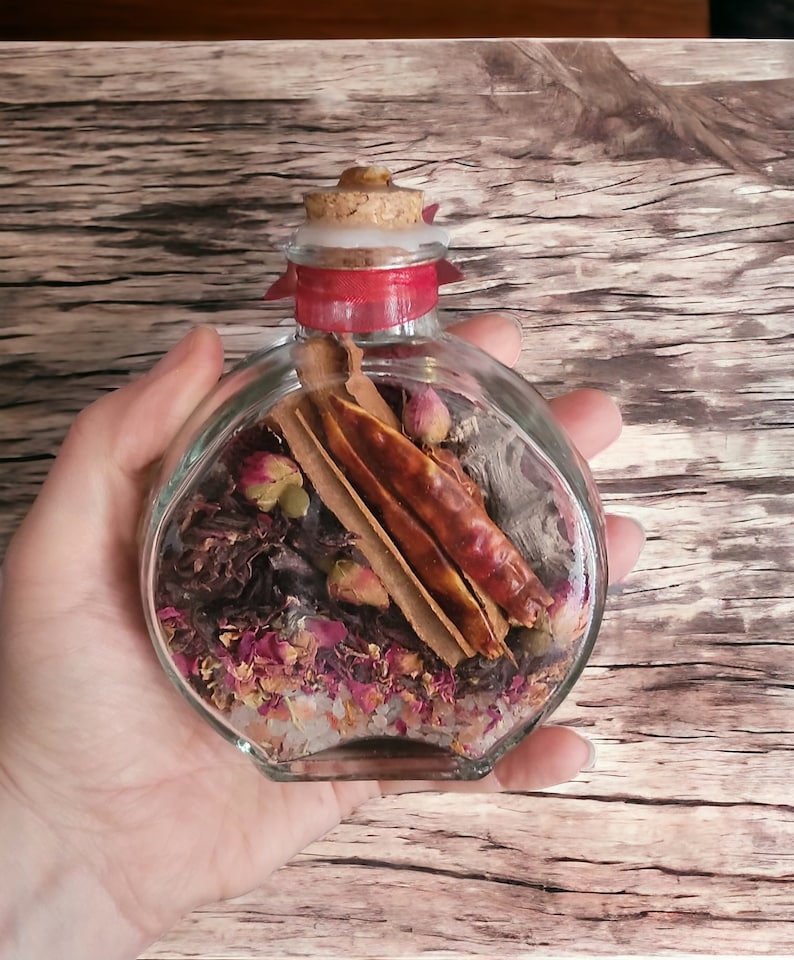 May include: A clear glass jar with a cork stopper and a red ribbon tied around the neck. The jar is filled with dried herbs, spices, and rose petals. The jar is sitting on a wooden surface.