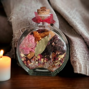 May include: A clear glass bottle with a cork stopper and a red ribbon tied around the neck. The bottle is filled with dried herbs, flowers, and spices, including bay leaves, cinnamon sticks, and rose petals.