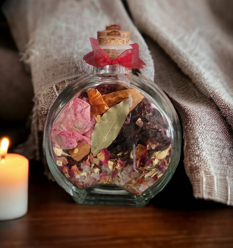 May include: A clear glass bottle with a cork stopper and a red ribbon tied around the neck. The bottle is filled with dried herbs, flowers, and spices, including bay leaves, cinnamon sticks, and rose petals.