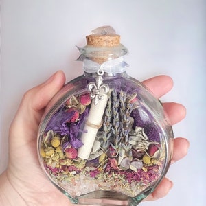May include: A clear, round glass bottle filled with dried flowers, herbs, and a rolled-up scroll. The bottle is topped with a cork and a clear crystal. A silver fleur-de-lis charm hangs from a ribbon tied around the neck.
