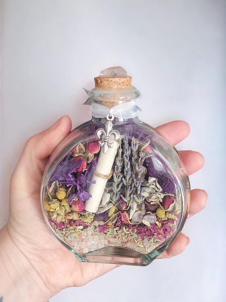 May include: A clear, round glass bottle filled with dried flowers, herbs, and a rolled-up scroll. The bottle is topped with a cork and a clear crystal. A silver fleur-de-lis charm hangs from a ribbon tied around the neck.