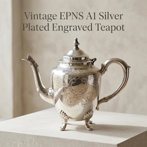 Puede incluir: Una tetera plateada con un patrón floral grabado, un asa curva, un pico y una tapa con un remate. La tetera se apoya sobre cuatro patas y presenta el texto Vintage EPNS A1 Silver Plated Engraved Teapot encima del objeto.