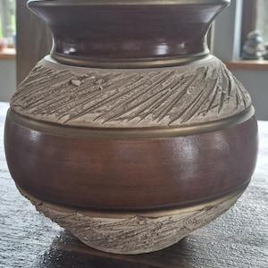 May include: A round ceramic vase featuring a smooth dark brown neck and midsection, contrasted by two bands of textured, light beige clay with diagonal carved markings. The vase has a wide, flared rim and a rounded body with a matte finish.