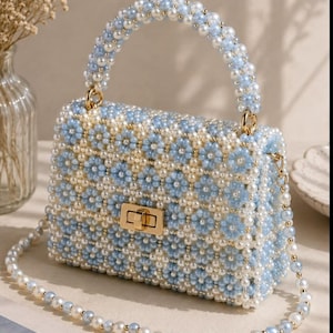 May include: A structured handbag crafted from light blue and white faux pearl beads arranged in a repeating floral pattern. The bag features a single top handle, a gold-toned metal turn-lock closure, and a detachable long shoulder strap made of beads.