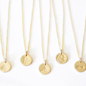 May include: A collection of gold-colored necklaces with delicate chains. Each necklace features a small, round pendant with an engraved design: an arrow, a wave, a dreamcatcher, a crescent moon with stars, and an upward-pointing arrow.