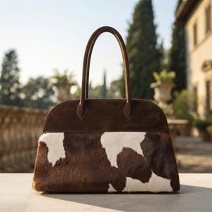 May include: A structured handbag featuring a dark brown suede upper section and a cowhide lower section with a brown and white spotted pattern. The bag includes two tall, thin brown handles attached to the top, with a curved silhouette.