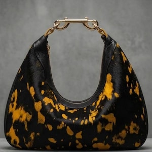 May include: A hobo-style handbag featuring a black and yellow cowhide pattern with a textured fur finish. The bag has a smooth black leather trim along the top opening and a prominent gold-toned metal hardware handle shaped like a carabiner.