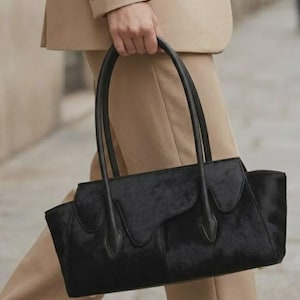 May include: A black handbag with a textured, hair-on-hide finish and a structured silhouette. The bag features two long, slender black handles and a distinctive flap closure with a scalloped, layered edge design across the front panel.