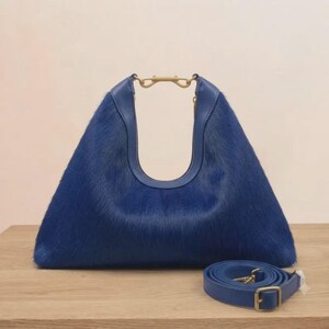 Könnte beinhalten: Eine leuchtend blaue Handtasche aus texturiertem Fellmaterial. Die Tasche hat eine markante dreieckige Silhouette mit einem glatten Lederbesatz entlang der oberen Öffnung und einem goldfarbenen Metallverschluss. Ein passender blauer Schulterriemen liegt bei.
