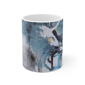 Puede incluir: Una taza cilíndrica de cerámica con un diseño abstracto que presenta capas texturizadas de pintura blanca, azul pizarra y negra. La superficie incluye pequeños detalles dorados en relieve y patrones de pinceladas visibles en el exterior.