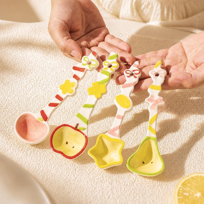 May include: Four ceramic spoons with white handles featuring colorful painted stripes, bows, and floral accents. The bowls of the spoons are shaped like a round circle, an apple, a star, and a pear, each in pastel pink, yellow, and light green.