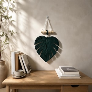 Peut inclure: Une suspension murale en macramé vert foncé en forme de feuille de monstera. L'ouvrage en fibre est suspendu à une tige en bois clair par un cordon blanc, avec une perle en bois unique au sommet central de la structure de la feuille.