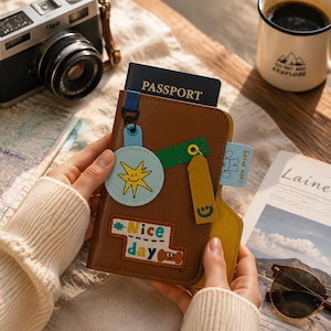 May include: A brown passport holder featuring a blue circular patch with a yellow sun graphic, a green fabric strap, a yellow rectangular tag with a blue smiley face, and a rectangular patch with the text Nice day and a brown dog illustration.
