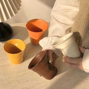 Puede incluir: Un conjunto de cuatro tazas de cerámica en colores naranja, amarillo, marrón y blanco roto. Cada taza presenta una textura arrugada o plegada distintiva en la sección central, similar a un vaso de papel, con un borde y base lisos.