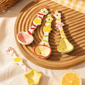 May include: Four ceramic spoons with decorative handles featuring colorful patterns and shapes. The spoons have unique bowl designs including a pink heart, a red-rimmed apple, a green-rimmed pear, and a yellow star, all with glossy finishes.