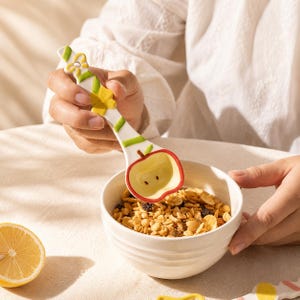 May include: A white ceramic spoon shaped like an apple with a red rim, a green stem, and a handle decorated with yellow flowers and green stripes, held over a white bowl filled with granola, accompanied by a sliced lemon on the table.