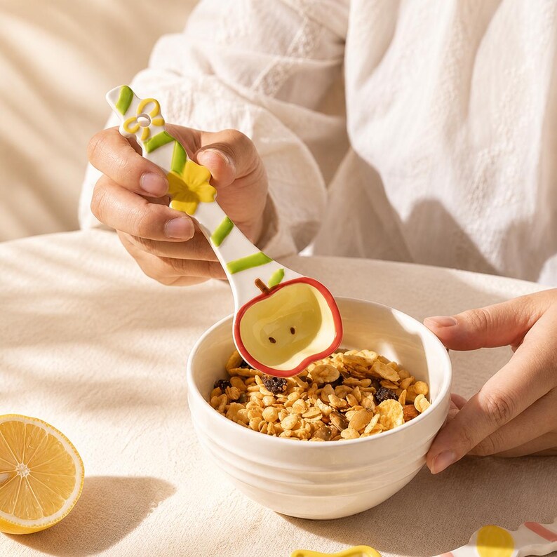 May include: A white ceramic spoon shaped like an apple with a red rim, a green stem, and a handle decorated with yellow flowers and green stripes, held over a white bowl filled with granola, accompanied by a sliced lemon on the table.