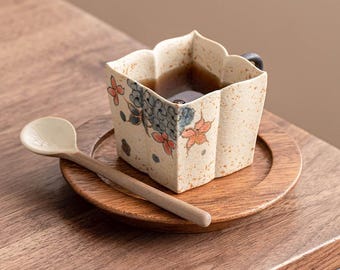 Könnte beinhalten: Eine quadratische Keramiktasse mit gewelltem Rand und gesprenkelter beiger Oberfläche, verziert mit handgemalten blauen und orangen Blumenmotiven. Die Tasse steht auf einer runden Holzuntertasse neben einem kleinen, hellen Keramiklöffel.