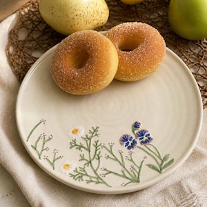 May include: A round, off-white ceramic plate featuring a hand-painted botanical design of small white daisies, purple and blue pansies, and green stems. Two golden-brown sugar-coated doughnuts rest on the surface of the plate.