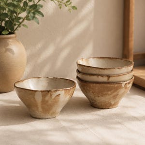 May include: A set of four ceramic bowls with an irregular, organic shape. The bowls feature a cream-coloured interior and a mottled, earthy brown exterior finish with visible brushstrokes and a textured, hand-crafted appearance.