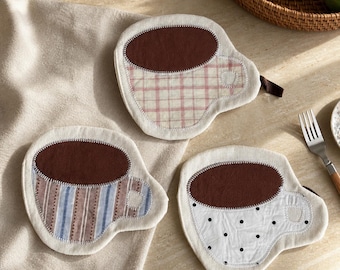 Könnte beinhalten: Drei Stoffuntersetzer in Form von Kaffeetassen, jeweils mit einer dunkelbraunen Filzoberseite. Ein Untersetzer hat ein rosa Karomuster, einer hat blaue und braune vertikale Streifen und einer ist weiß mit kleinen schwarzen Punkten.