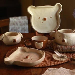 May include: A collection of cream and brown ceramic tableware featuring bear-shaped designs. The set includes two plates, two bowls with ear-like protrusions, and a brown mug with a white bear face relief, all with small brown noses.