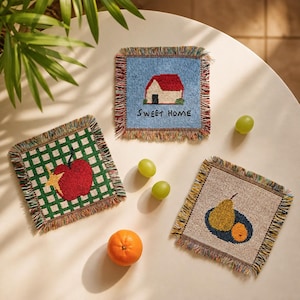 May include: Three square woven coasters with fringed edges. One features a red house on a blue background with the text SWEET HOME. Another displays a red apple on a green and white checkered pattern. The third shows a yellow pear and orange.