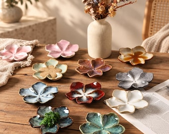 Könnte beinhalten: Eine Sammlung kleiner, blumenförmiger Keramikschalen in verschiedenen Farben wie Rosa, Blau, Rot, Weiß, Braun und Grün. Jede Schale hat fünf abgerundete Blütenblätter und eine glasierte Oberfläche, angeordnet auf einer hölzernen Oberfläche.