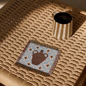 May include: A square woven coaster featuring a light blue and white geometric star pattern with a central brown amphora motif and frayed edges, accompanied by a ceramic mug with vertical brown and white stripes and a dark interior.