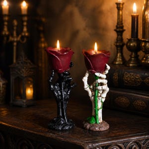 May include: Two candle holders shaped like skeletal hands, one black and one white, each grasping a green stem topped with a deep red rose-shaped candle. Both candles are lit, casting a warm glow from the center of the rose petals.