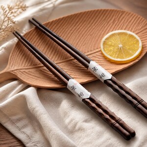 May include: A pair of dark brown wooden chopsticks with a textured, scalloped handle design. Each chopstick features a white paper band printed with black Japanese characters. The chopsticks are resting on a light brown wooden leaf-shaped tray.