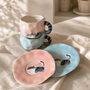 Puede incluir: Dos tazas de cerámica y dos platillos a juego en rosa pastel y azul claro. Cada taza presenta una cara de gato atigrado gris pintada a mano y un asa de cola rayada. Los platillos muestran un gato gris pintado sobre un borde ondulado.