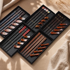 May include: Five black rectangular gift boxes containing sets of chopsticks and matching rests. The sets feature various designs including blue patterns, floral motifs, red accents, and natural wood finishes with distinct shapes for the rests.