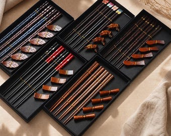 Könnte beinhalten: Fünf schwarze rechteckige Geschenkboxen mit Essstäbchen-Sets und passenden Ablagen. Die Sets zeigen verschiedene Designs, darunter blaue Muster, florale Motive, rote Akzente und Ausführungen in Naturholz mit unterschiedlichen Formen.