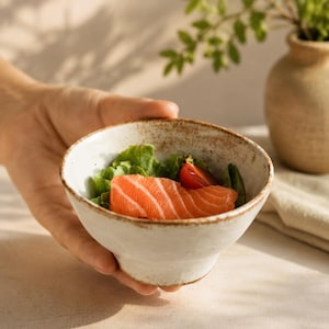 May include: A small, round, off-white ceramic bowl with a speckled brown rim. The bowl contains a piece of bright orange raw salmon fillet, a single red cherry tomato, and green lettuce leaves, showcasing a rustic, textured finish.