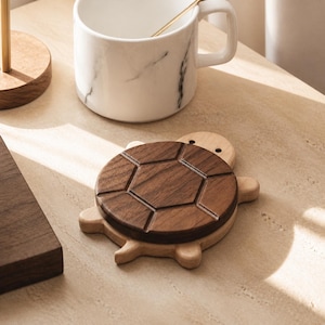 May include: A turtle-shaped coaster featuring a light-colored wooden base and a dark brown walnut wood shell with a carved hexagonal pattern. The coaster has a smooth finish and is designed with four small legs and a head extending from the body.