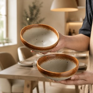 Könnte beinhalten: Zwei Keramikschalen mit gesprenkeltem, cremeweißem Innenraum und einem breiten, texturierten rostfarbenen Rand. Die Schalen haben eine abgerundete Form und eine glatte Oberfläche, die einen deutlichen Kontrast zwischen der hellen Mitte und dem dunklen Rand zeigt.