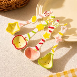 May include: A set of four ceramic spoons with white handles featuring colorful painted patterns of bows, flowers, and stripes. Each spoon has a unique bowl shape including a star, an apple, a heart, and a pear, with vibrant yellow, red, and green accents.