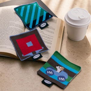 Peut inclure: Trois dessous de verre carrés en tricot avec des motifs distincts : un à rayures diagonales vertes et bleues, un avec un carré rouge et un centre bleu sur fond marron, et un avec un motif de voiture bleue. Chaque pièce porte une étiquette Wishmore.