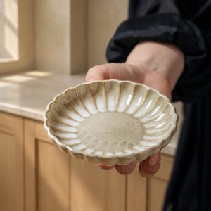 Könnte beinhalten: Eine kleine, runde Keramikschale mit einem gewellten, blütenblattartigen Rand. Das Innere weist eine strukturierte, hellbeige Mitte auf, die in ein weiß glasiertes, geripptes Muster übergeht, das zum geschwungenen, geriffelten Rand strahlt.