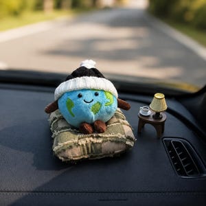 Peut inclure: Une peluche en forme de globe terrestre au visage souriant, portant un bonnet en tricot noir et blanc, posée sur un coussin à carreaux verts et beiges. À côté se trouve une table miniature marron avec une petite lampe et une tasse blanche.