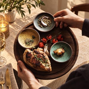 Peut inclure: Une planche de service ronde en bois foncé portant un blanc de poulet grillé aux herbes, trois petits bols en céramique contenant des épices, des tomates cerises rôties et des gousses d'ail, sur une surface texturée avec un verre de vin blanc.