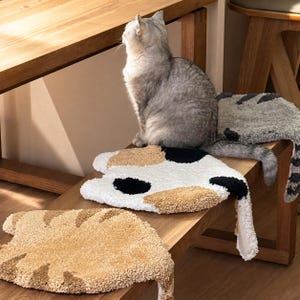 Può includere: Tre tappeti trapuntati a forma di gatto con motivi distinti: un tappeto beige con strisce più scure, uno bianco con macchie nere e beige e uno grigio con strisce scure, tutti caratterizzati da superfici testurizzate e code pendenti.