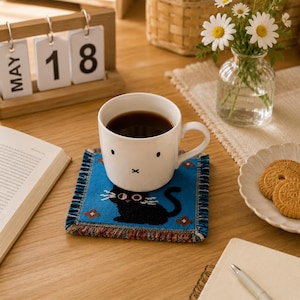 May include: A white ceramic mug with a simple black face graphic sits on a square blue woven coaster featuring a black cat illustration with white whiskers and small floral accents. The coaster has a fringed edge with multicoloured threads.